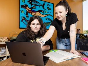 One person talks to another person who’s sitting at a desk while pointing at a laptop